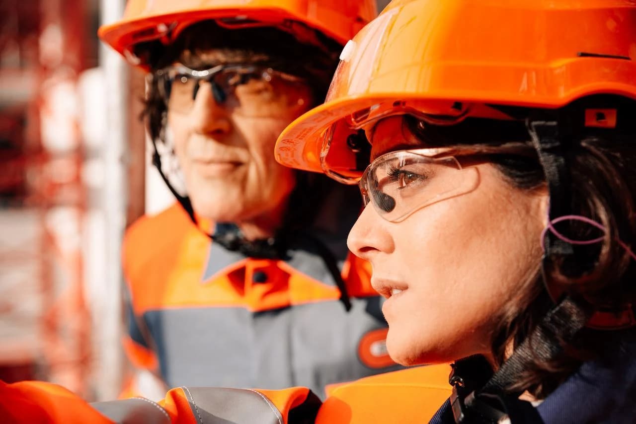 Deux travailleurs portent des casques de sécurité orange vif et des lunettes de protection. La femme au premier plan, en profil et vêtue d'un uniforme de travail gris et orange, regarde attentivement sur le côté.