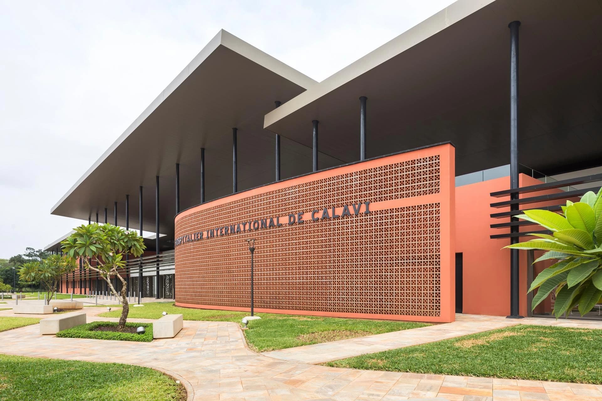 Façade moderne et incurvée de l'Hôpital International de Calavi en briques ajourées orange, surmontée d'un large toit gris clair soutenu par des piliers noirs, entourée de pelouses vertes et d'allées pavées.