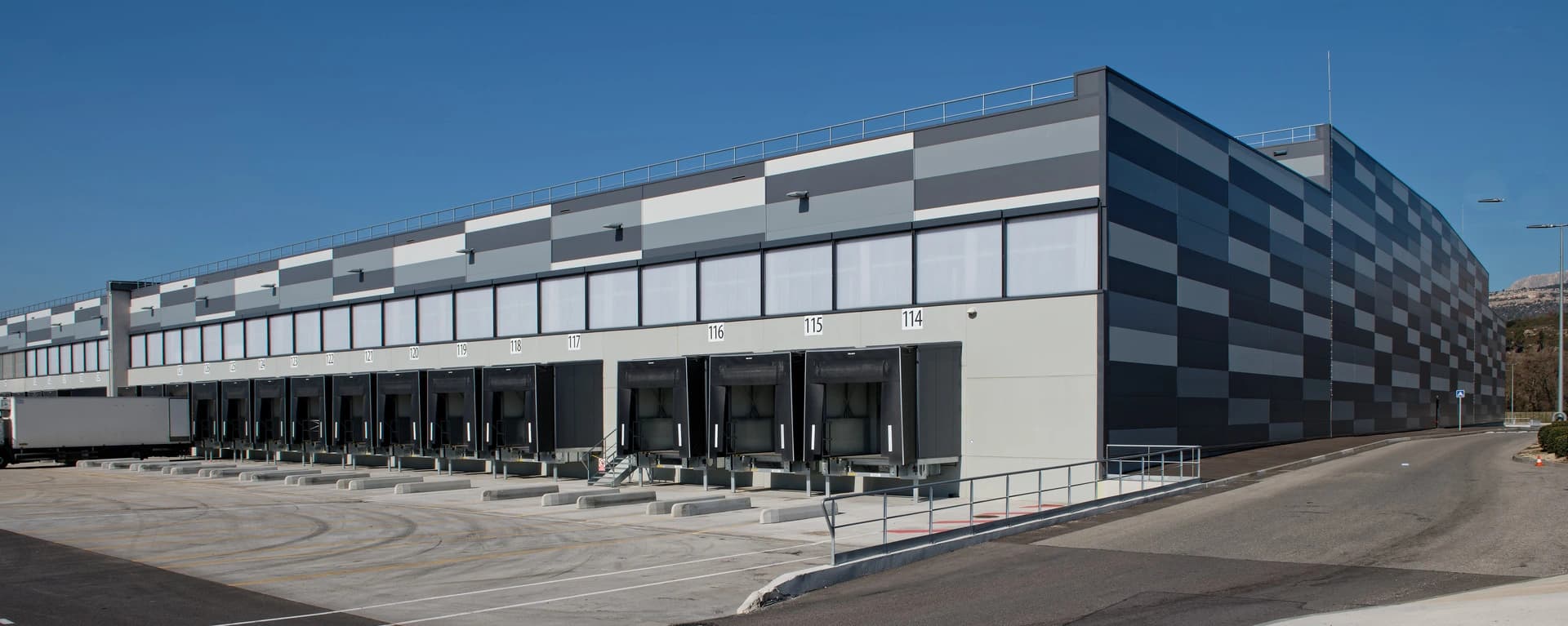 Grand bâtiment logistique moderne avec une longue rangée de quais de chargement numérotés, sa façade grise et blanche à motifs sous un ciel bleu clair, avec un camion blanc garé et des montagnes en arrière-plan.