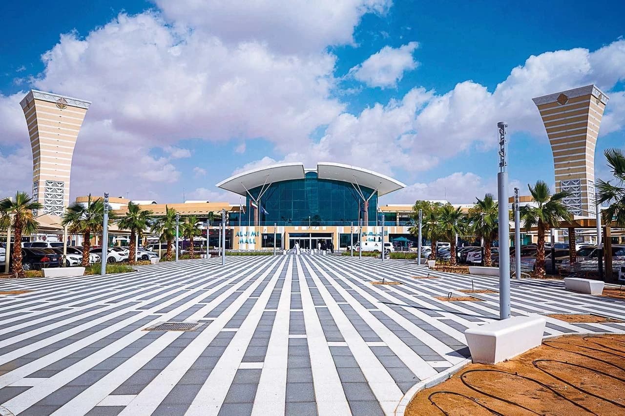 Vue extérieure du centre commercial Al Jimi Mall, un bâtiment moderne avec une façade vitrée et un toit incurvé, flanqué de hautes tours et de palmiers, avec une place pavée à rayures au premier plan sous un ciel bleu.