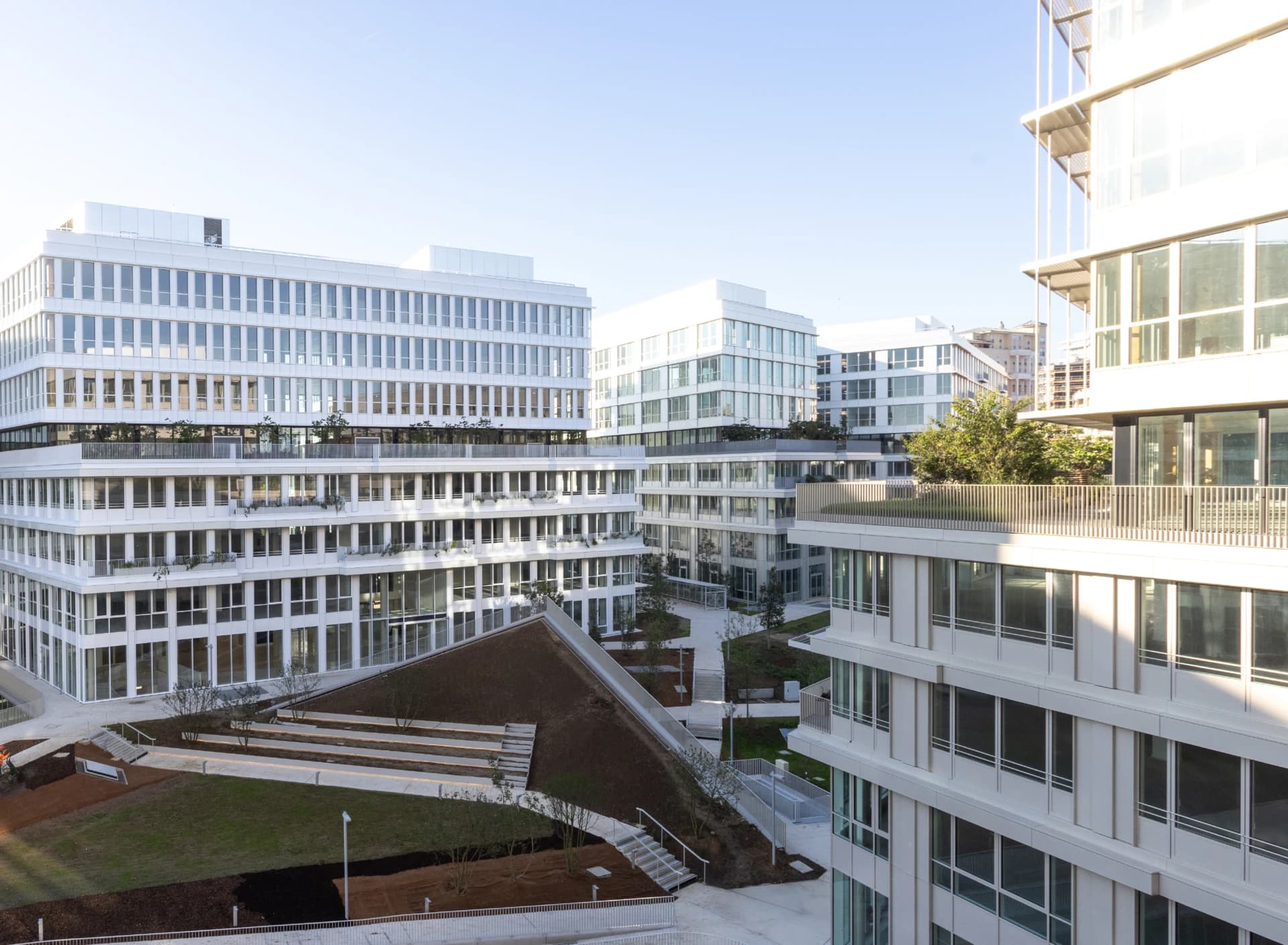 Vue d'un complexe moderne lumineux composé de bâtiments blancs aux nombreuses fenêtres, entourant une cour paysagée avec des pelouses en terrasses, des allées et une toiture végétalisée en pente.