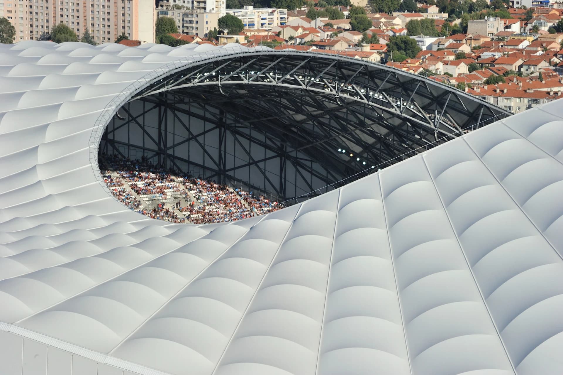 Vue plongeante sur le toit blanc ondulé d'un stade moderne, avec une ouverture révélant une section des gradins remplie de nombreux spectateurs et la structure métallique sombre. En arrière-plan, une ville dense de toits rouges et d'immeubles s'étend sous un ciel clair.