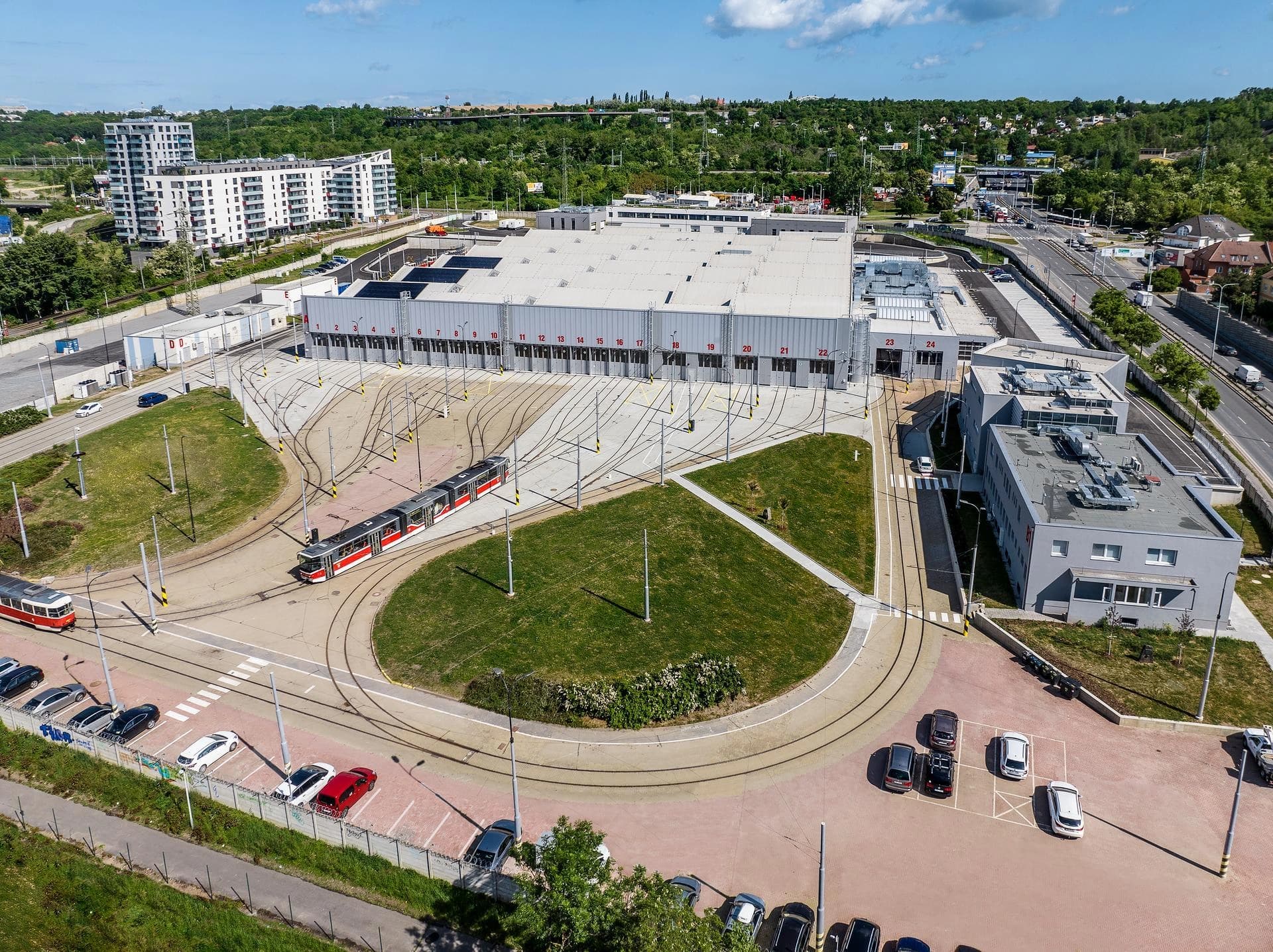 Vue aérienne d'un grand dépôt de tramway, montrant un vaste bâtiment gris de maintenance avec 24 baies et un réseau complexe de voies ferrées. Plusieurs trams rouges et blancs circulent sur les voies, dans un environnement urbain avec des immeubles, des routes et des espaces verts sous un ciel bleu et nuageux.
