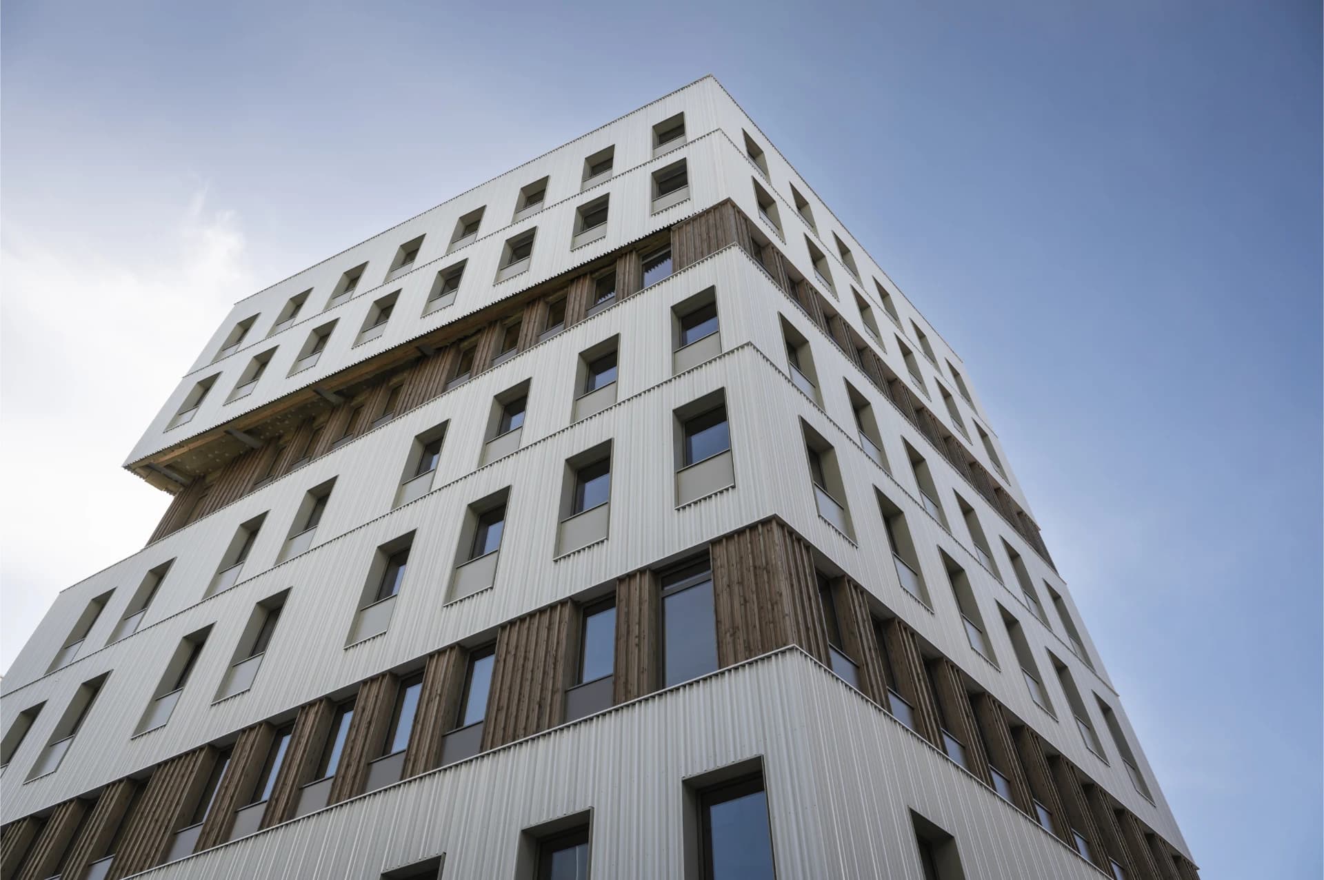 Un immeuble moderne et anguleux, avec des façades alternant des panneaux métalliques ondulés blancs et un revêtement en bois sombre, s'élève contre un ciel bleu clair.