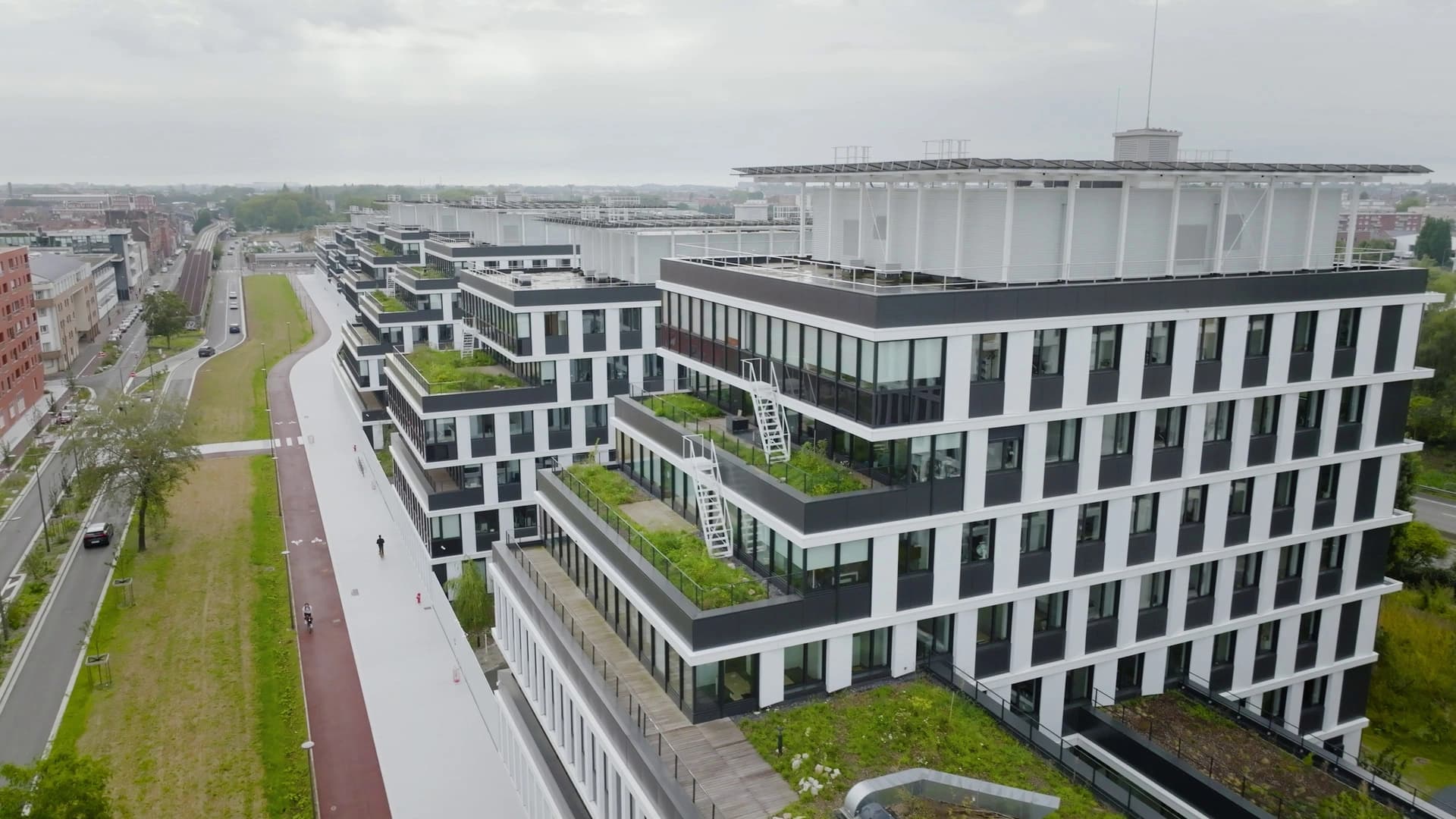 Un complexe de bâtiments modernes avec des façades noires et blanches, doté de toits verts en terrasses, s'étend le long d'une longue allée piétonne et cyclable. Ces structures au design durable sont entourées de verdure et d'une zone urbaine sous un ciel couvert.