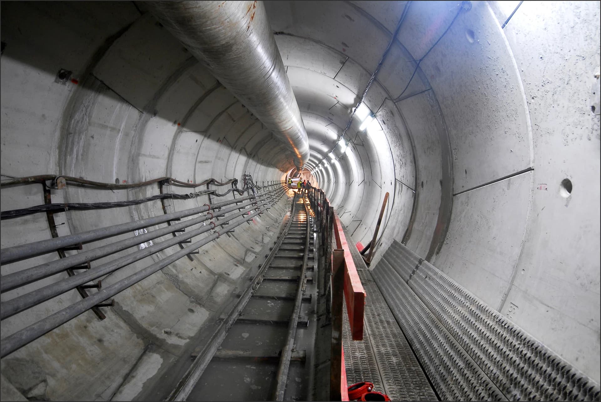 Vue en perspective d'un tunnel en béton brut en construction, éclairé par des luminaires et s'étendant vers des ouvriers au loin. Des tuyaux, câbles et une grande conduite courent le long des parois, tandis qu'un chemin de fer étroit et un trottoir grillagé avec une main courante rouge occupent le sol.