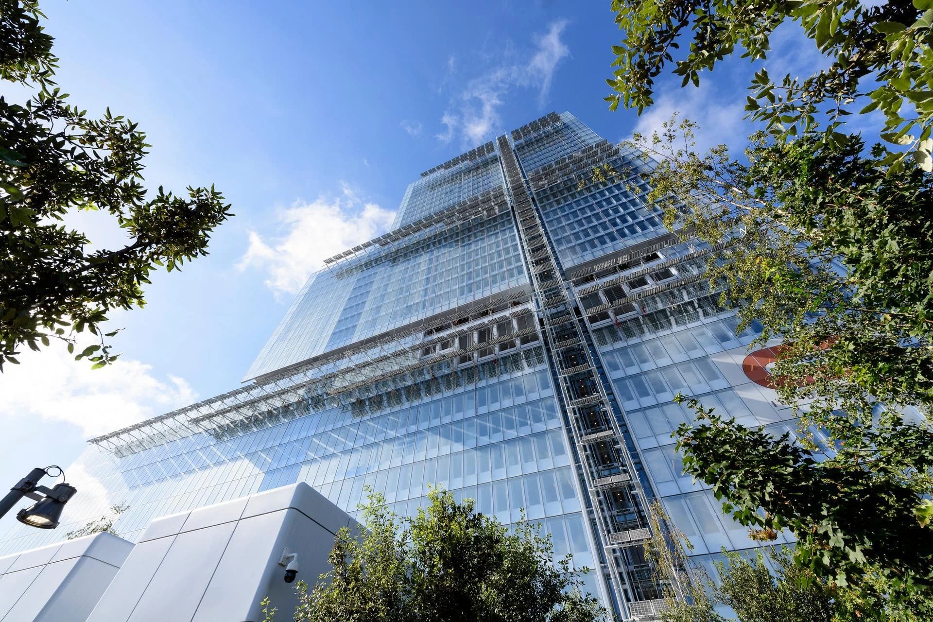 Un gratte-ciel moderne et vitré s'élève vers un ciel bleu parsemé de nuages blancs, encadré par des feuillages verts luxuriants. La façade réfléchissante de l'immeuble capte la lumière du soleil, créant des teintes bleues et grises.