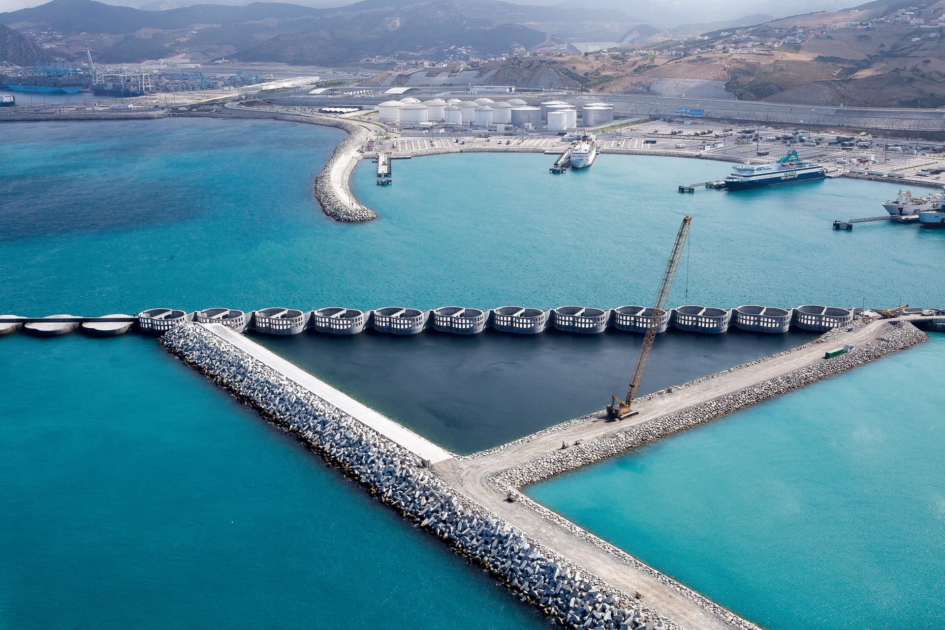 Vue aérienne d'un grand port industriel où des eaux turquoise et bleu foncé bordent des brise-lames en béton et des structures circulaires, avec une grue visible au premier plan. À l'arrière-plan, des navires, des réservoirs de stockage blancs et des montagnes côtières s'étendent sous un ciel clair.