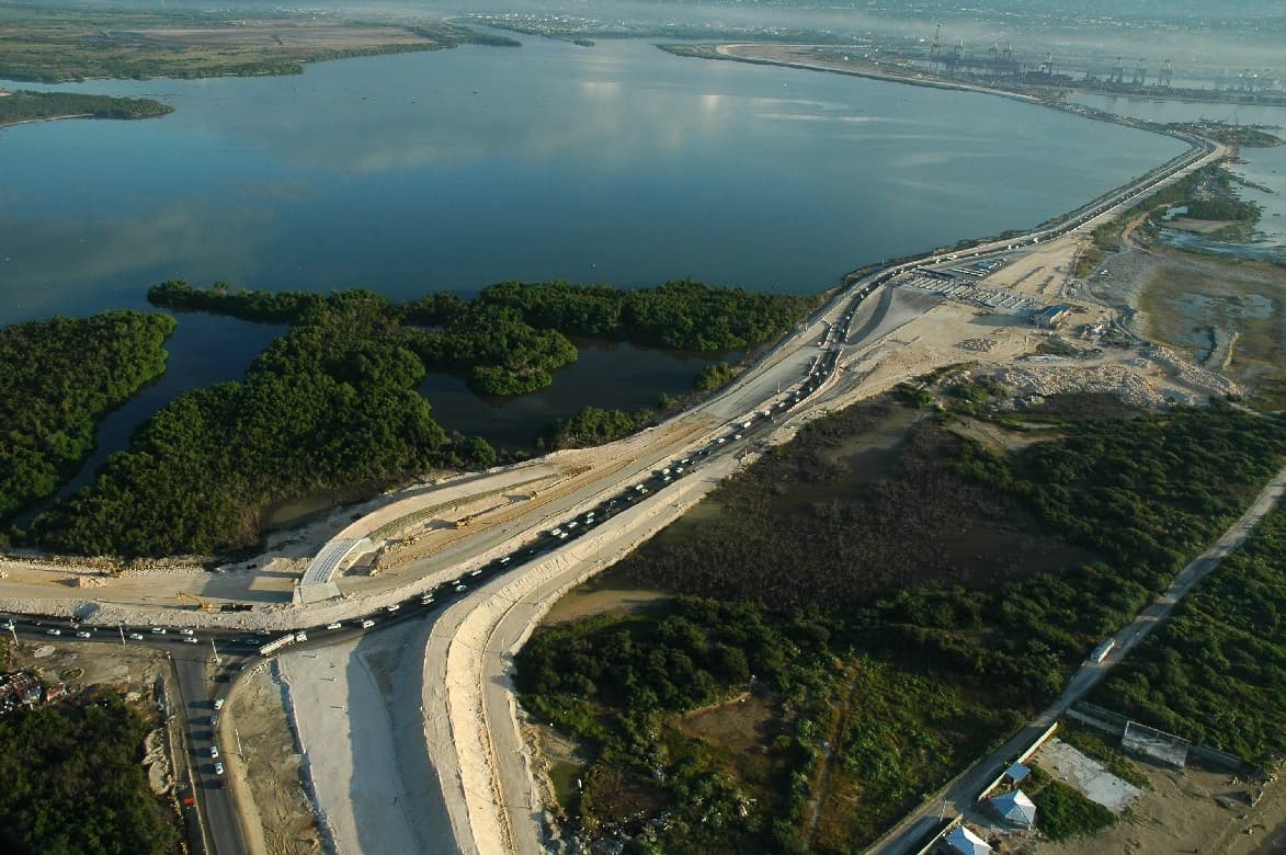 Vue aérienne d'une autoroute côtière moderne avec des voitures, longeant une vaste étendue d'eau calme bordée de mangroves vertes, avec des zones de construction et un port industriel à l'horizon.