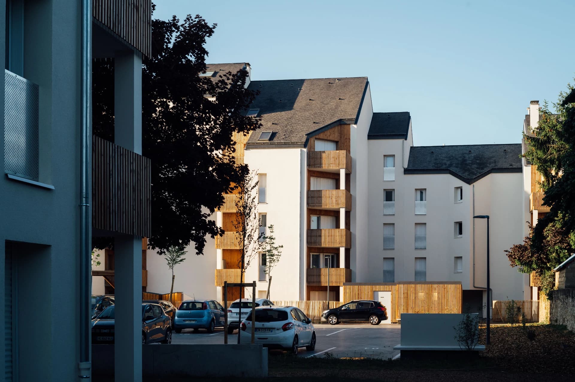 Un complexe résidentiel moderne présente des façades blanches, des balcons à lattes de bois clair et des toits sombres, avec plusieurs voitures garées dans un parking ensoleillé, le tout sous un ciel bleu clair.