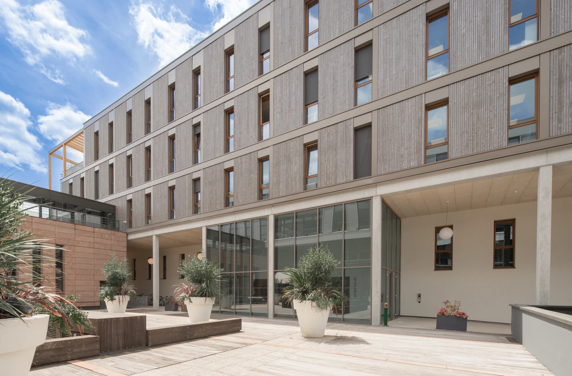 Un bâtiment moderne de plusieurs étages avec un bardage en bois clair et de grandes fenêtres à cadres bruns domine une cour ensoleillée avec terrasse en bois et grands pots de plantes vertes, sous un ciel bleu nuageux.