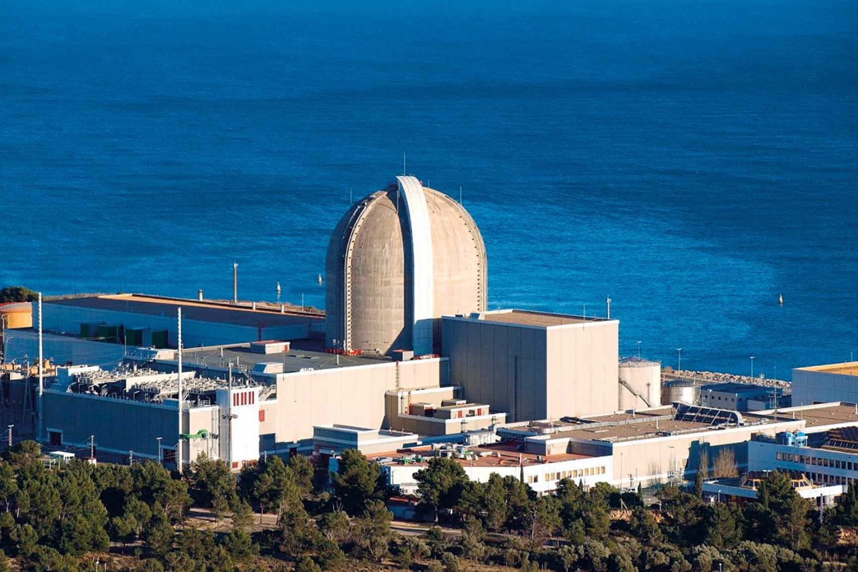 Une centrale nucléaire imposante avec un grand dôme gris clair se dresse au milieu de bâtiments industriels et de végétation verdoyante, face à une vaste étendue d'eau bleue profonde.