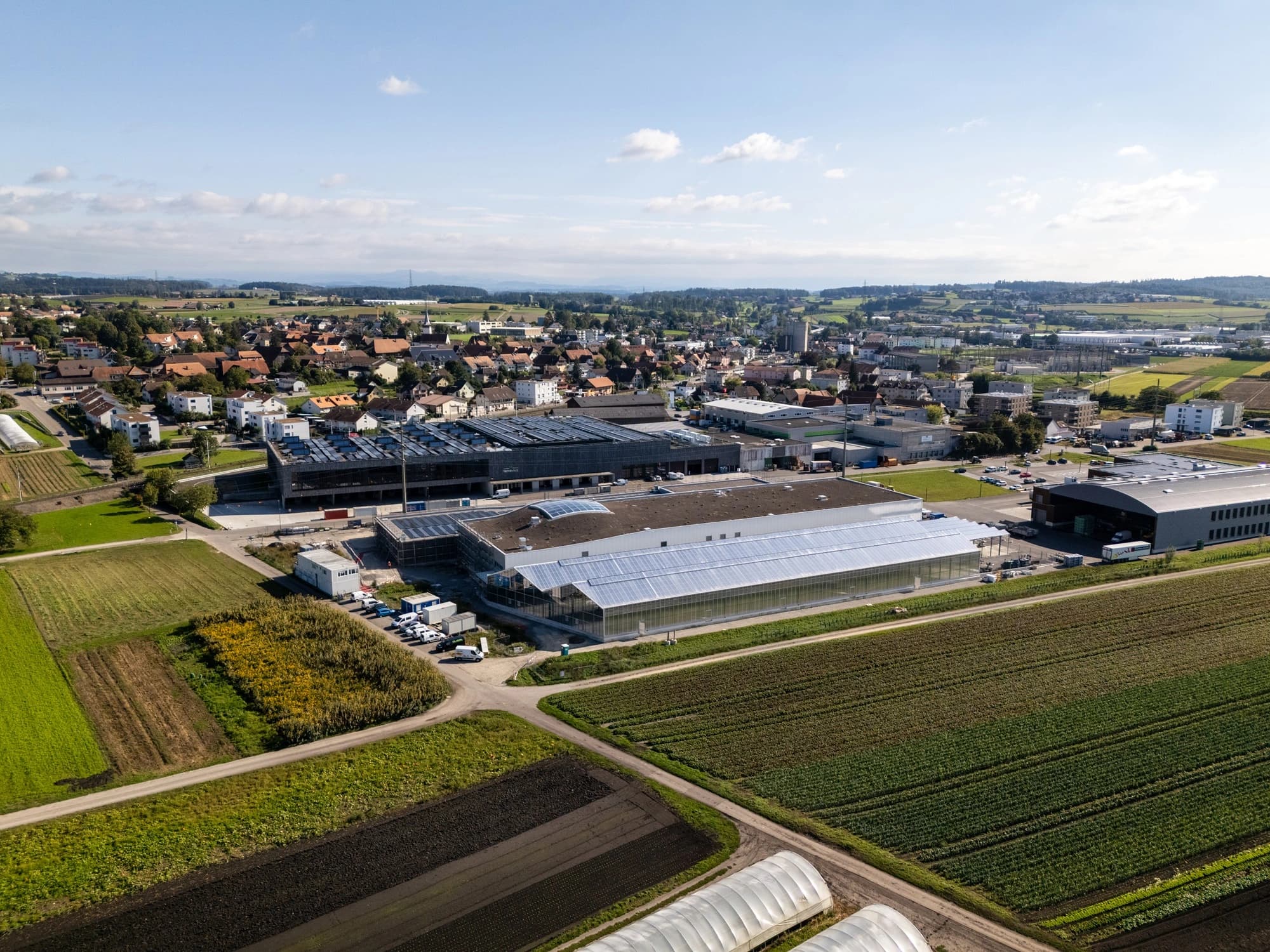 Vue aérienne d'un complexe agro-industriel moderne avec de grandes serres et un bâtiment équipé de panneaux solaires, bordé de champs agricoles verts et de parcelles de terre brune, avec une ville résidentielle en arrière-plan sous un ciel bleu parsemé de nuages blancs.