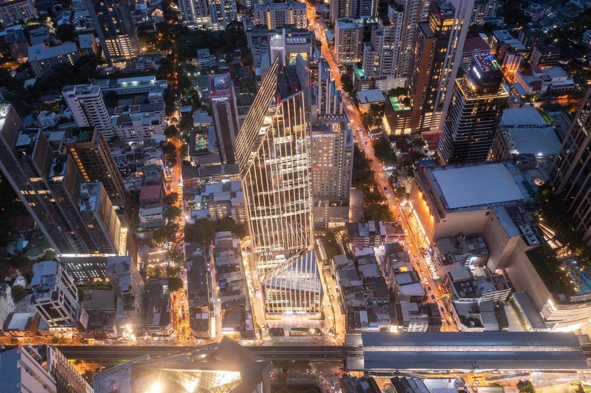 Vue aérienne nocturne d’un gratte-ciel moderne illuminé au cœur d’un quartier d’affaires dynamique, entouré d’immeubles et d’avenues urbaines éclairées.