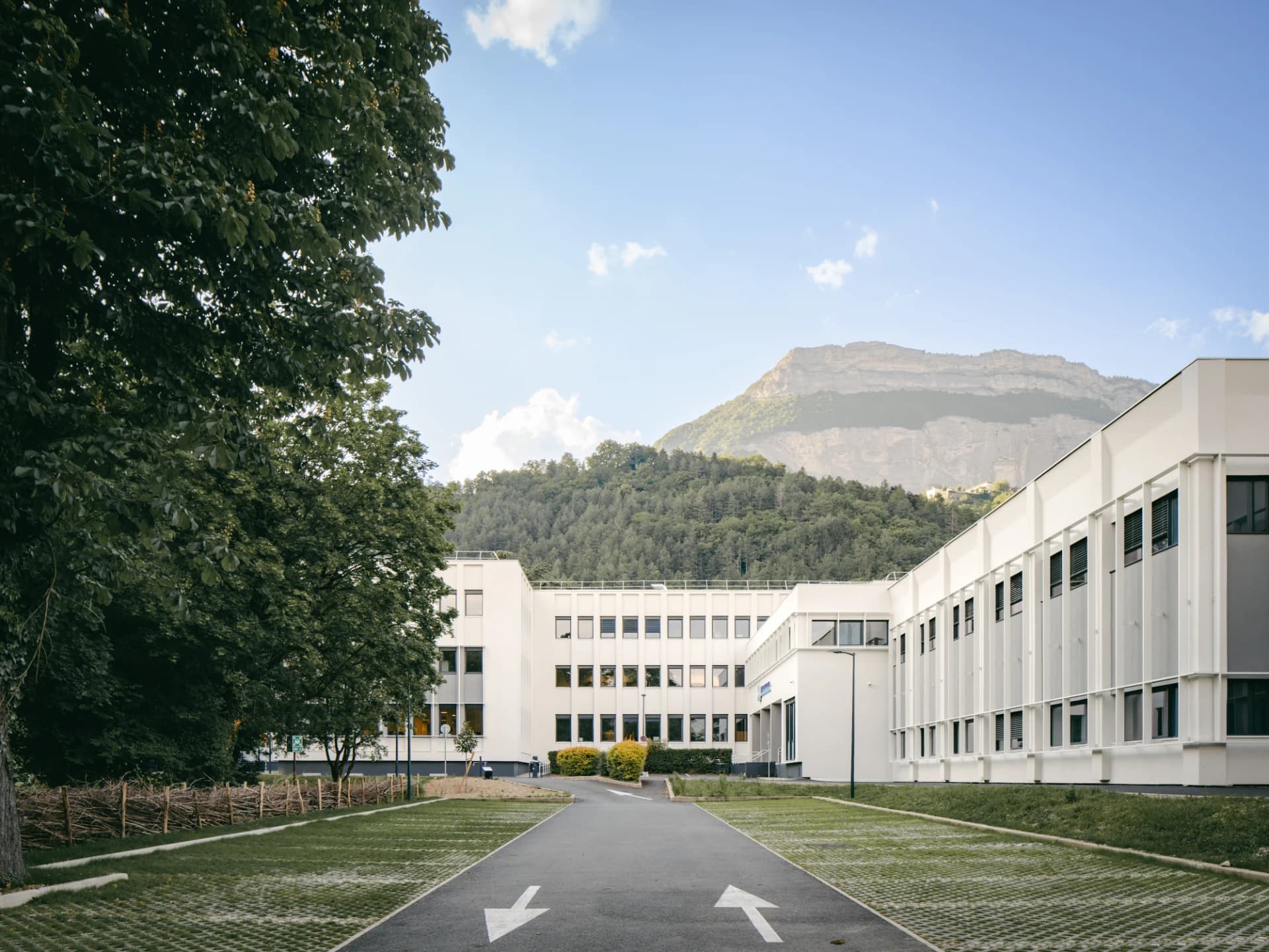 Un moderne bâtiment blanc avec de grandes fenêtres sombres se dresse devant une imposante montagne boisée surmontée d'une paroi rocheuse, avec un chemin d'accès bitumé et des places de parking engazonnées au premier plan, sous un ciel bleu.