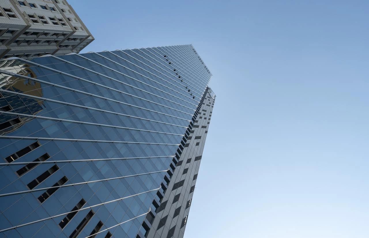 Un gratte-ciel moderne avec une façade en verre bleu-gris, photographié d'en bas, reflétant un autre bâtiment sur sa surface, sous un ciel bleu clair.