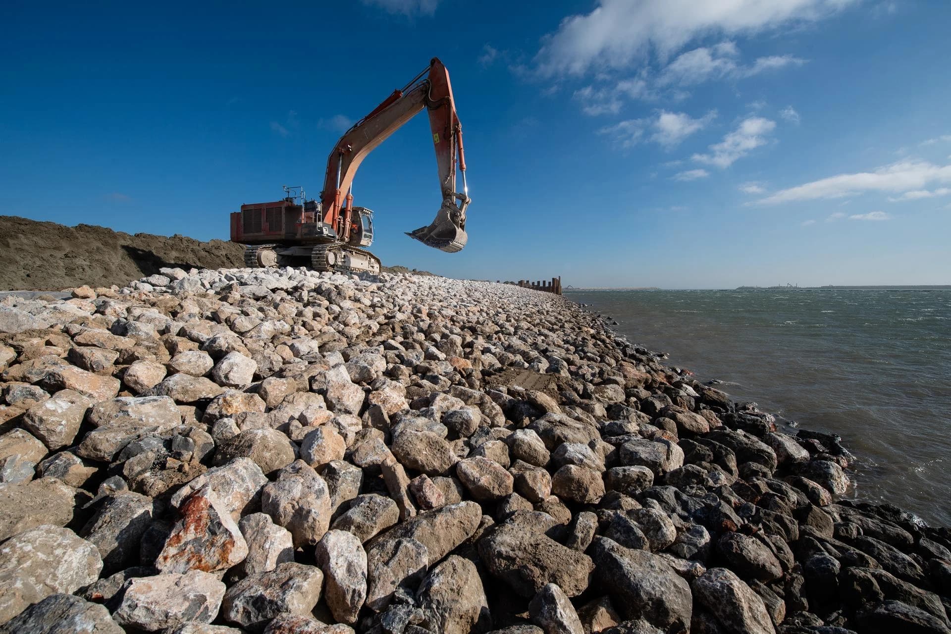 Un grand excavateur orange repose sur une longue barrière rocheuse fraîchement construite, longeant une étendue d'eau sombre et agitée sous un ciel bleu vif parsemé de nuages blancs, illustrant un projet d'ingénierie côtière.