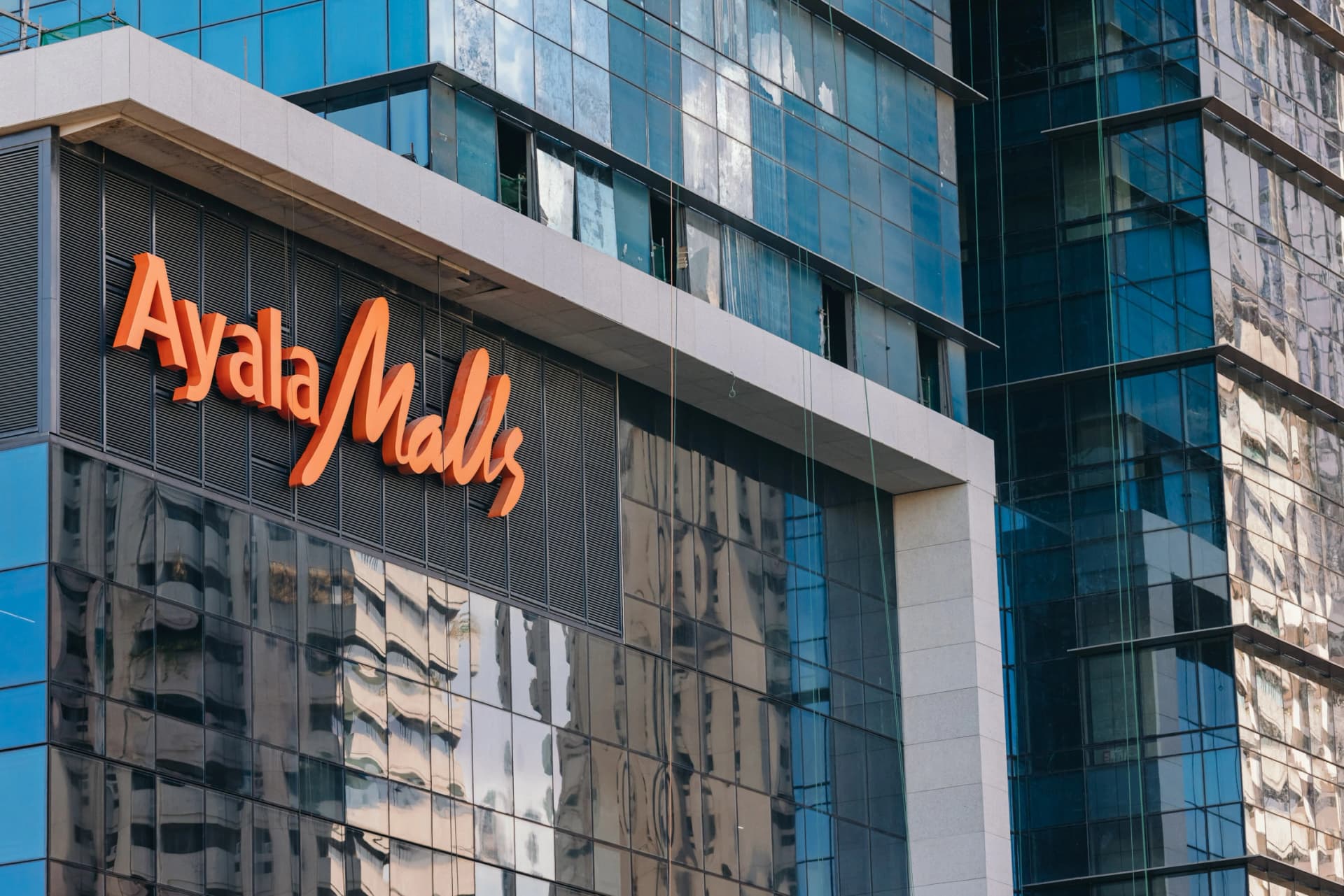 Un grand panneau orange vif "Ayala Malls" est visible sur la façade d'un immeuble moderne composé de panneaux de verre bleus et réfléchissants, et de quelques sections de béton gris clair.
